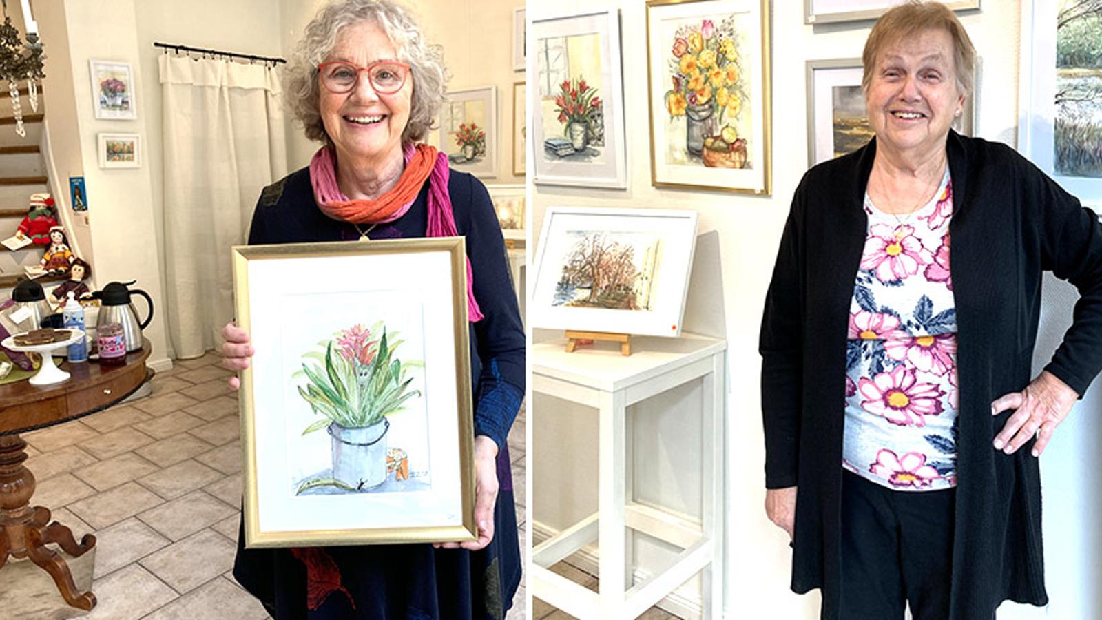 Två glada damer på vernissage. Damen till vänster håller upp en tavla som föreställer en vas med blommor. I bakgrunden hänger det tavlor på väggarna.