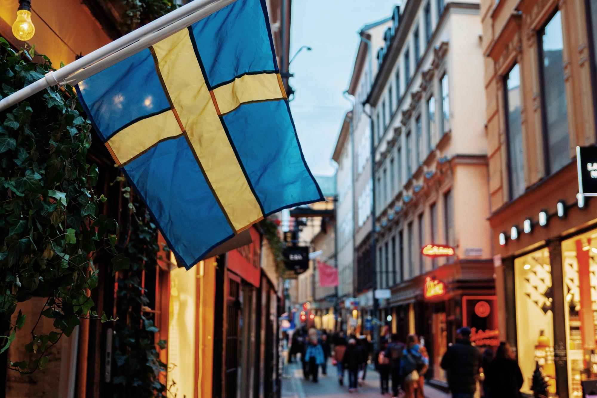 En butik, på en gata, med en svensk flagga som hänger vid entrén. I bakgrunden syns människor som går på gatan.