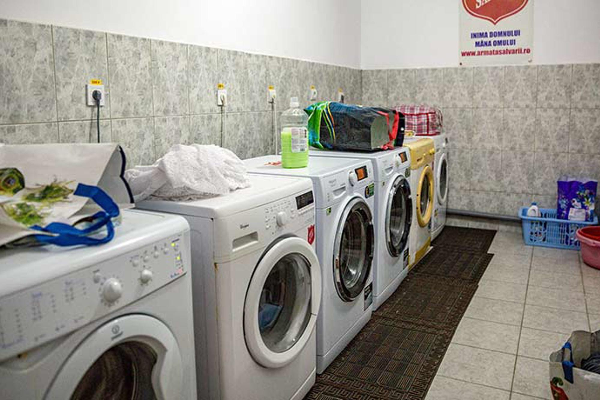 En tvättstuga med 6 tvättmaskiner som står på rad. I tvättstugan ligger det kassar, tvättmedel och sköljmedel. 