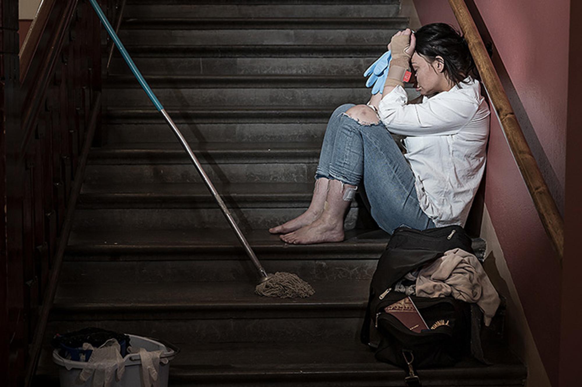En ung och utmattad kvinna som sitter i en trappuppgång med ansiktet i händerna. Hon har ett plåster på ena foten och hennes ena handled är inlindad i bandage. Runtom henne står det städutrutsning.