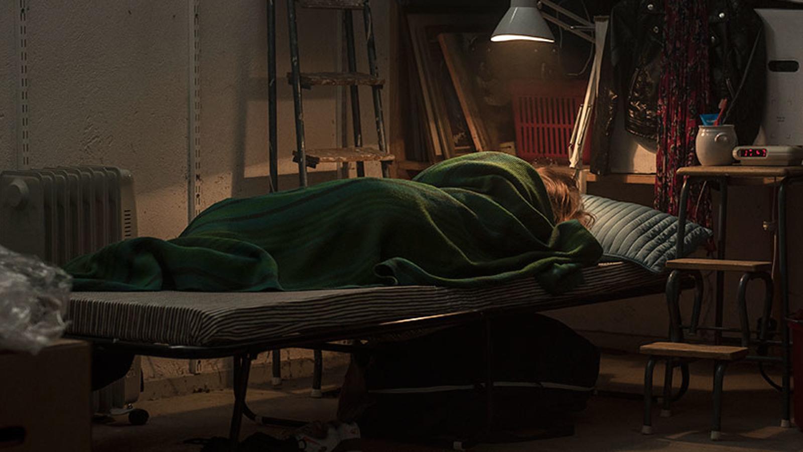 En kvinna som ligger inlindad i en filt på en säng. I rummet lyser en lampa ovanför sängen, och bakgrunden är stökig.