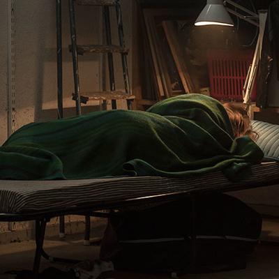 En kvinna som ligger inlindad i en filt på en säng. I rummet lyser en lampa ovanför sängen, och bakgrunden är stökig.
