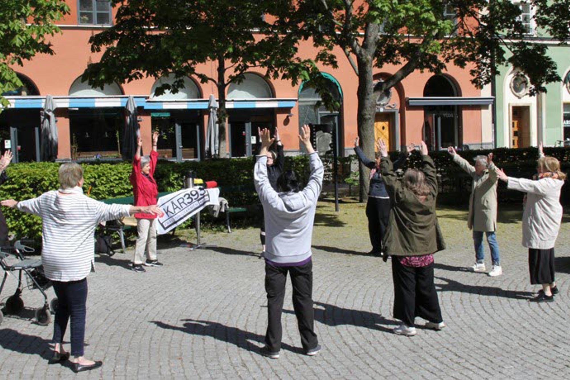 Äldre deltagare på utegympan som arrangeras av Kår 393 på Greta Garbos torg på Södermalm i Stockholm. Deltagarna sträcker armarna upp i luften.