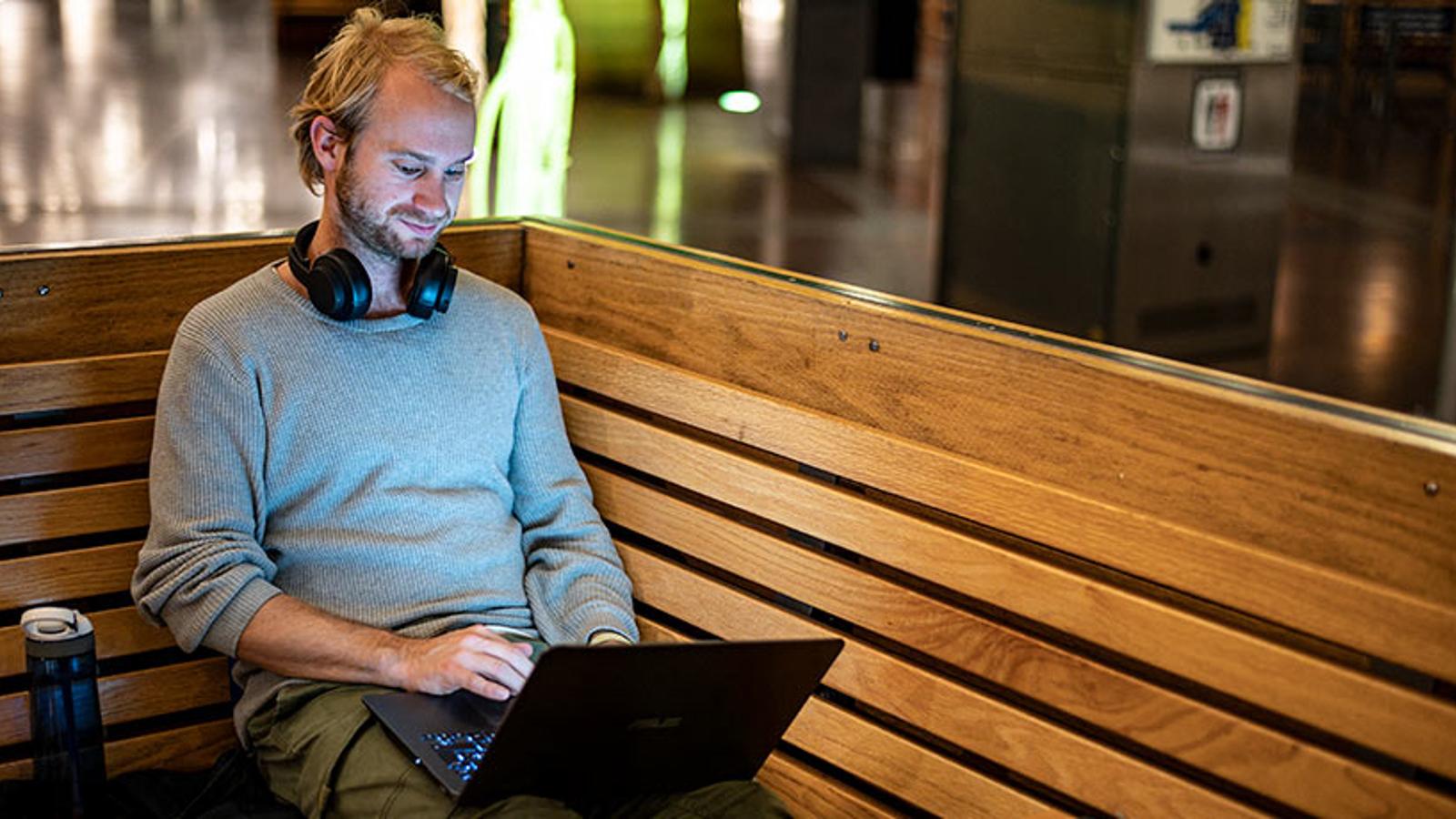 En man, Olav, som sitter på en bänk med en dator i knät. Han har ett par hörlurar runt halsen och bredvid honom står det en vattenflaska.
