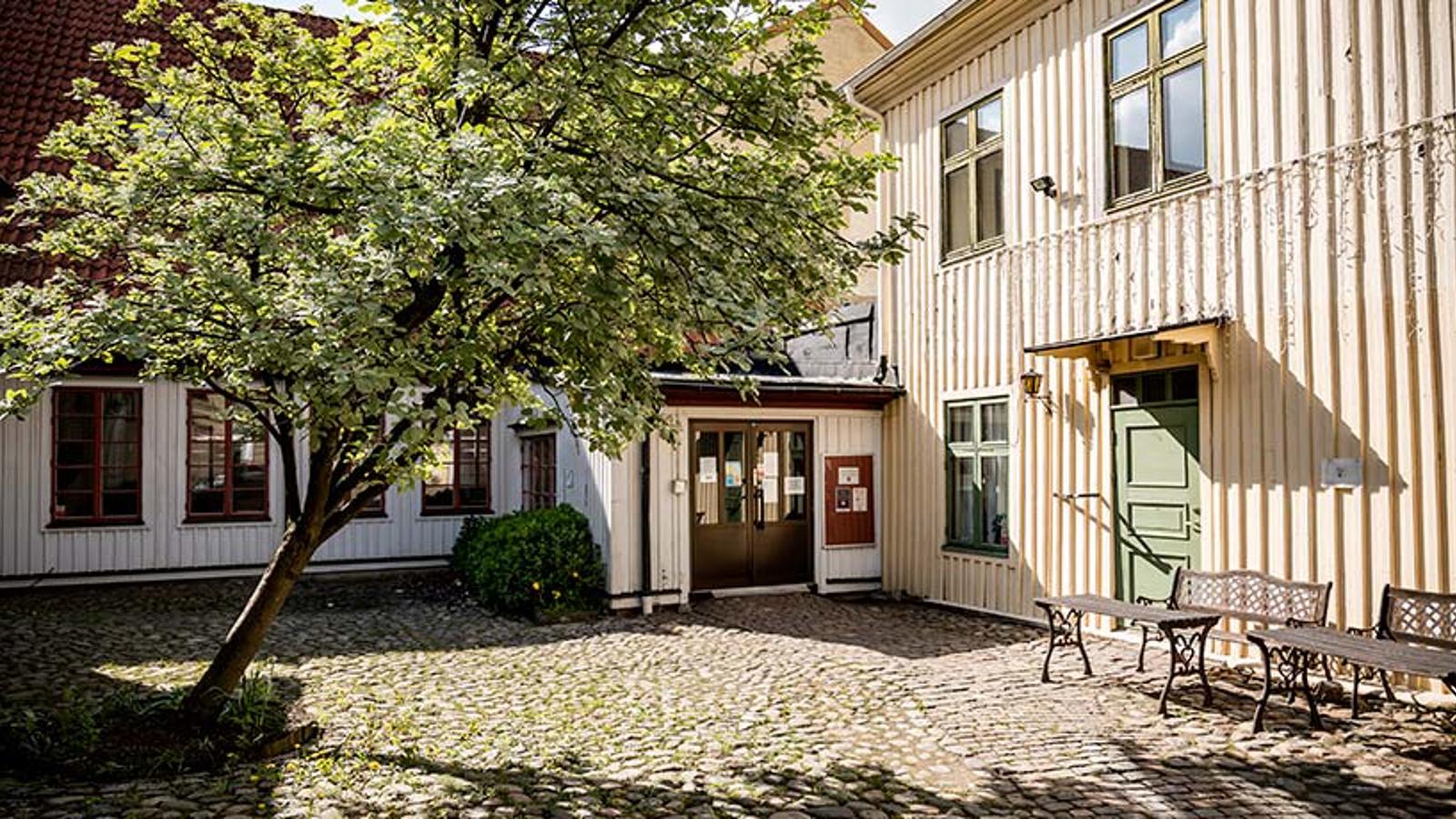 Centrumkåren Haga Mölndals fasad. På gården vid entrén står ett träd, till vänster, och till höger står två bänkar med bord.