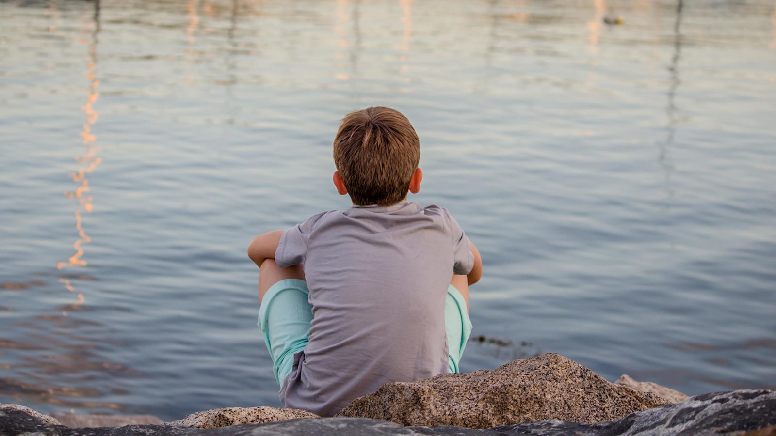 En pojke som sitter med ryggen mot kameran och tittar ut mot vattnet.