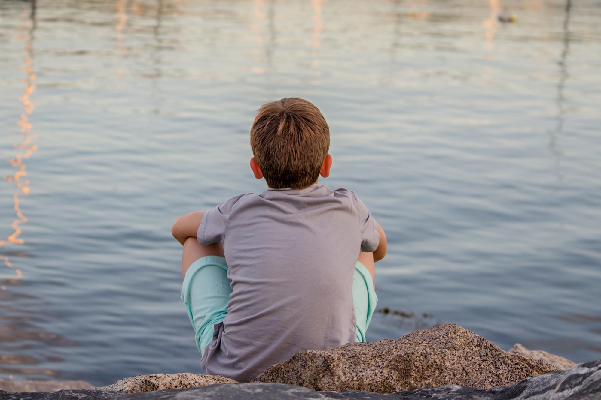 En pojke som sitter med ryggen mot kameran och tittar ut mot vattnet.