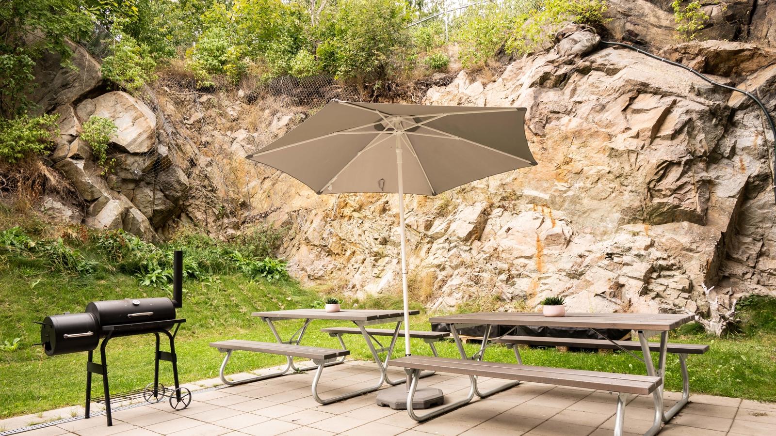 Uteplats med en grill, uppfällt parasoll och bänkar vid ett bord.