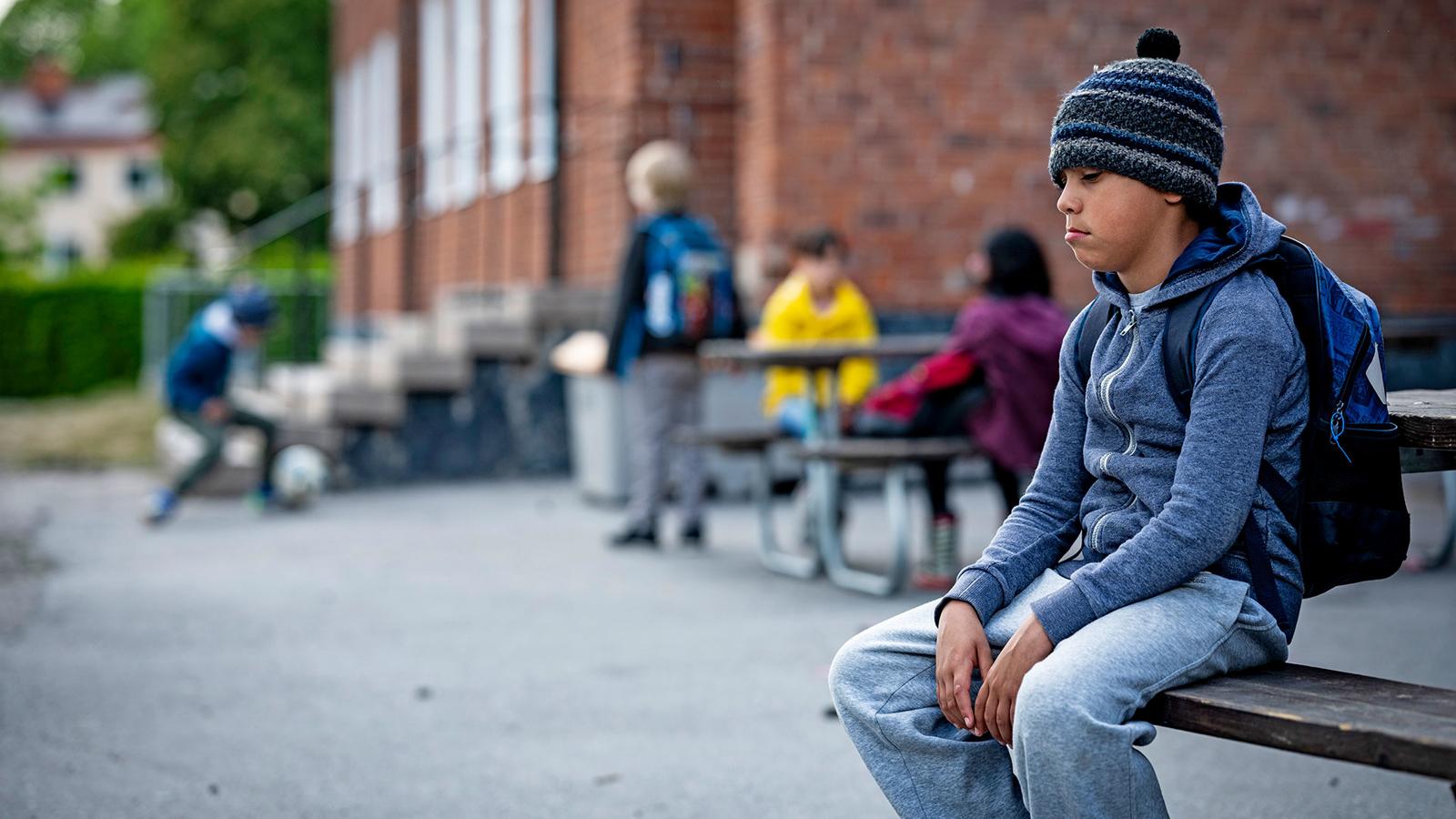 En ung pojke sitter ensam på en bänk på en skolgård. I bakgrunden syns fyra andra barn som leker.