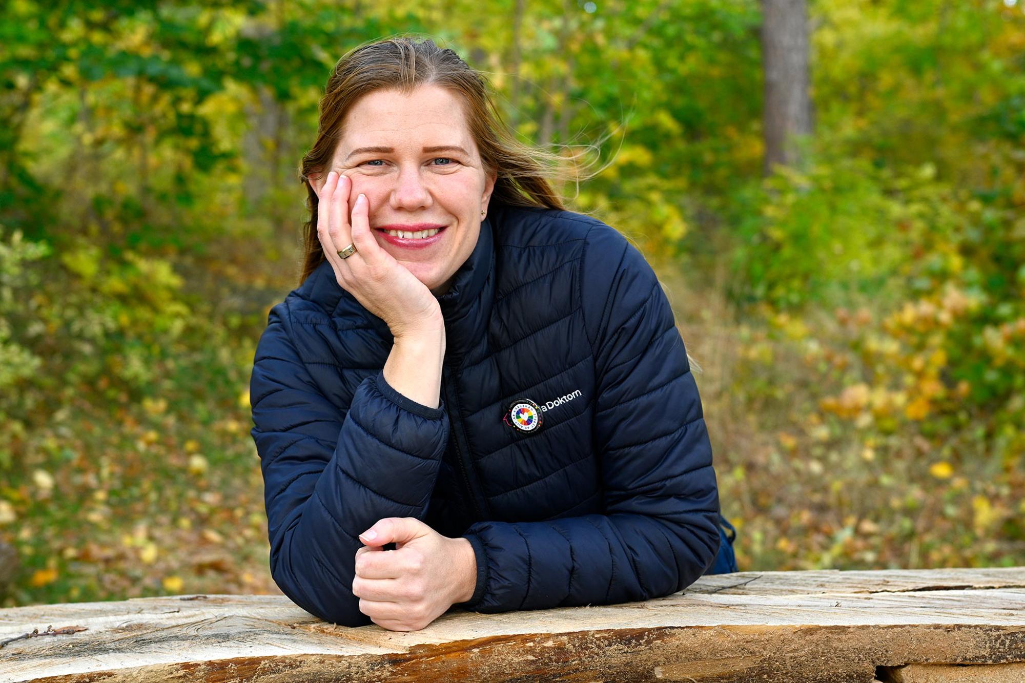 Helen lutar sig mot ett trästaket och tittar leende in i kameran, i bakgrunden syns skogen.