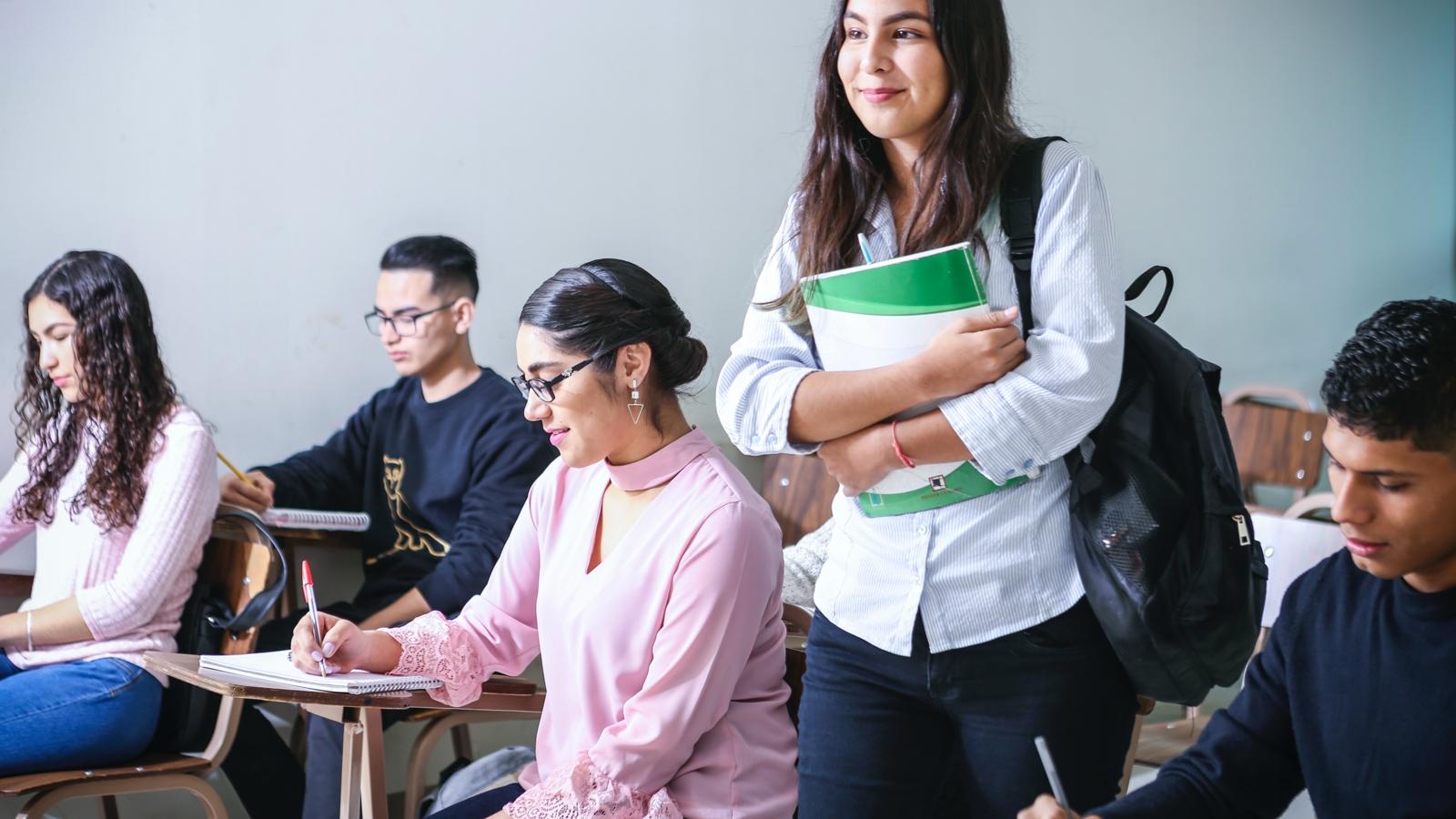 En ung kvinna med ryggsäck som går in i ett klassrum och håller i ett anteckningsblock. I klassrummet sitter det elever vid bänkar som sitter och skriver.
