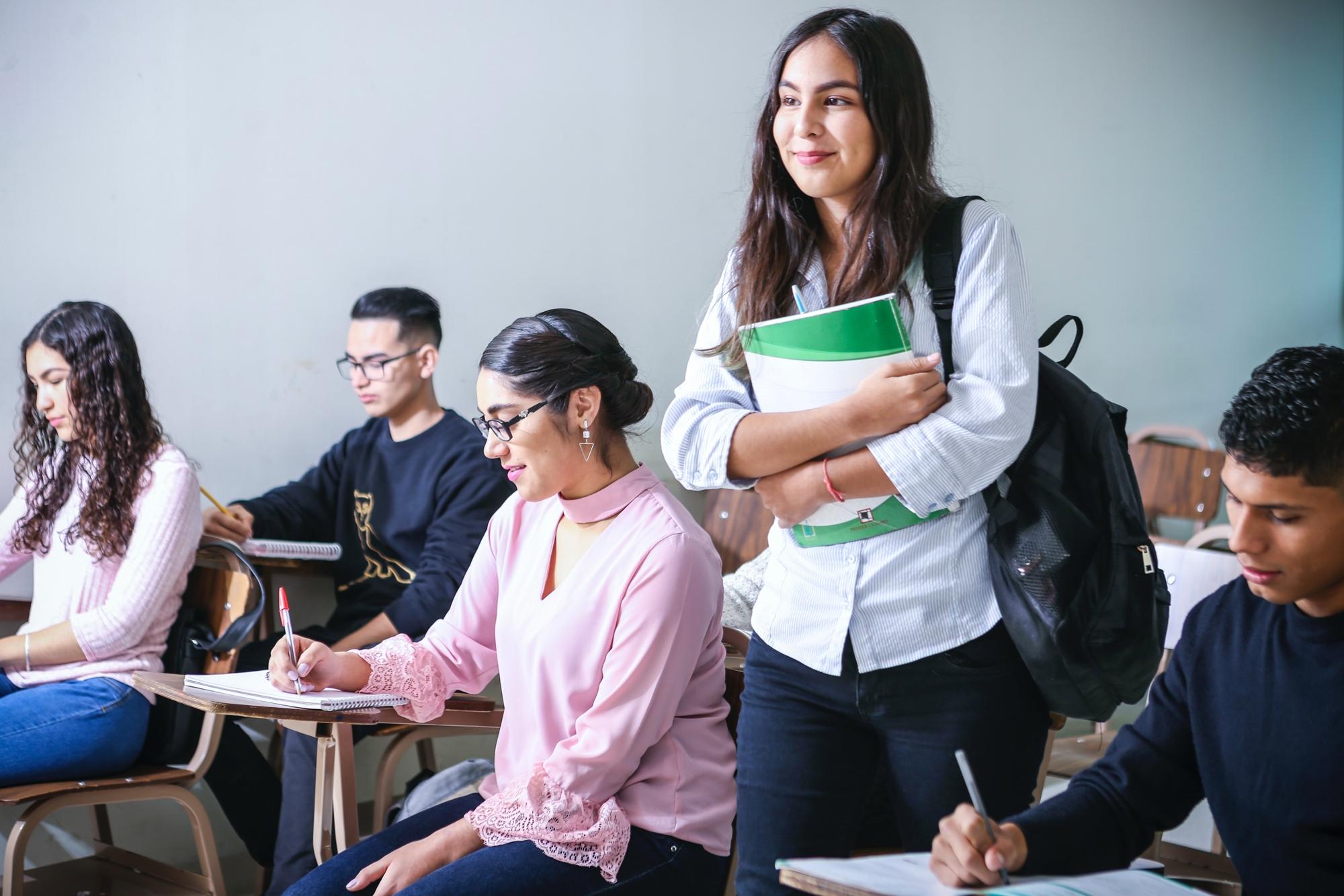 En ung kvinna med ryggsäck som går in i ett klassrum och håller i ett anteckningsblock. I klassrummet sitter det elever vid bänkar som sitter och skriver.