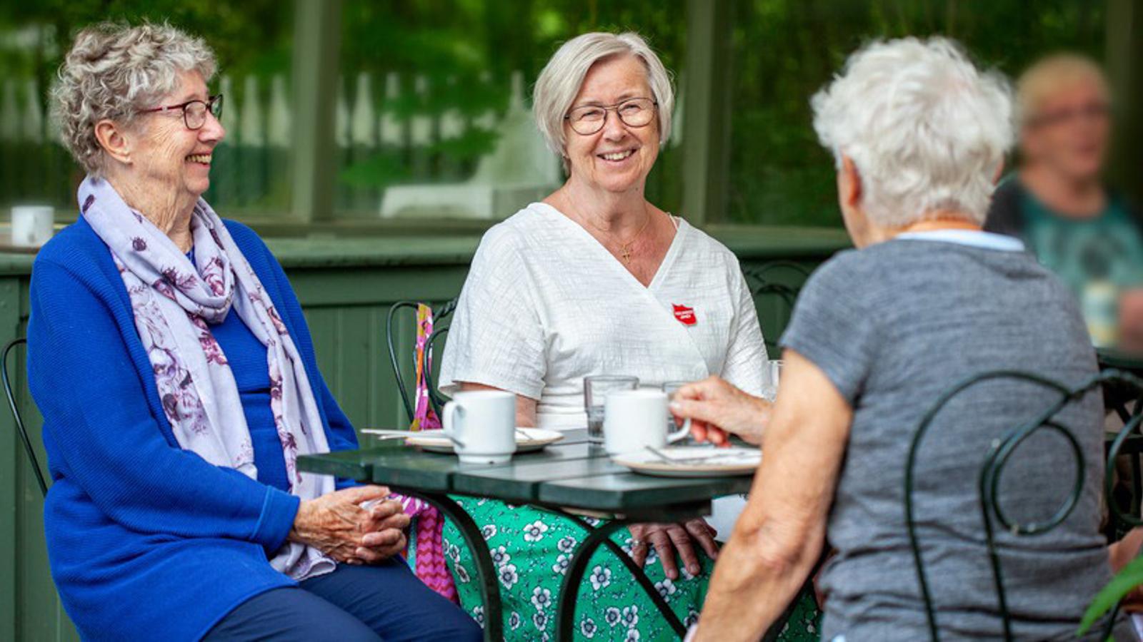 Två glada äldre kvinnor sitter vid ett cafébord tillsammans med en kvinna som har Frälsningsarméns sköld på tröjan.