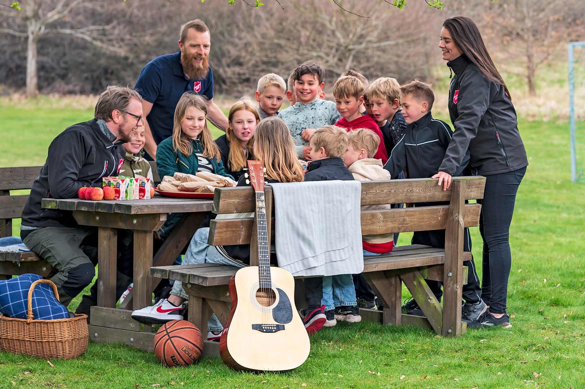 Tre personer från Frälsningsarmén tillsammans med glada barn runt ett picknickbord med frukt, dricka och mackor. Framför bordet ligger det en basketboll och en korg med en filt i. Det står även en gitarr lutad emot bordet.
