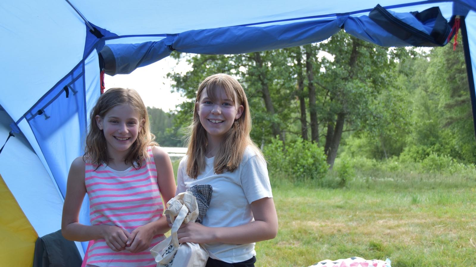 Agnes och Signe är med på sitt första scoutläger. De står tillsammans vid ett tält ute i naturen och ler mot kameran.
