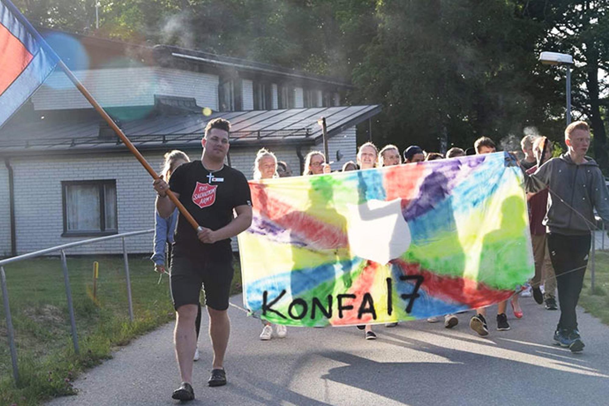 Längst fram går det en man från Frälsningsarmén som håller i en flagga. Bakom honom går ett gäng konfirmander som håller i ett färgglatt skynke med texten "Konfa 17".