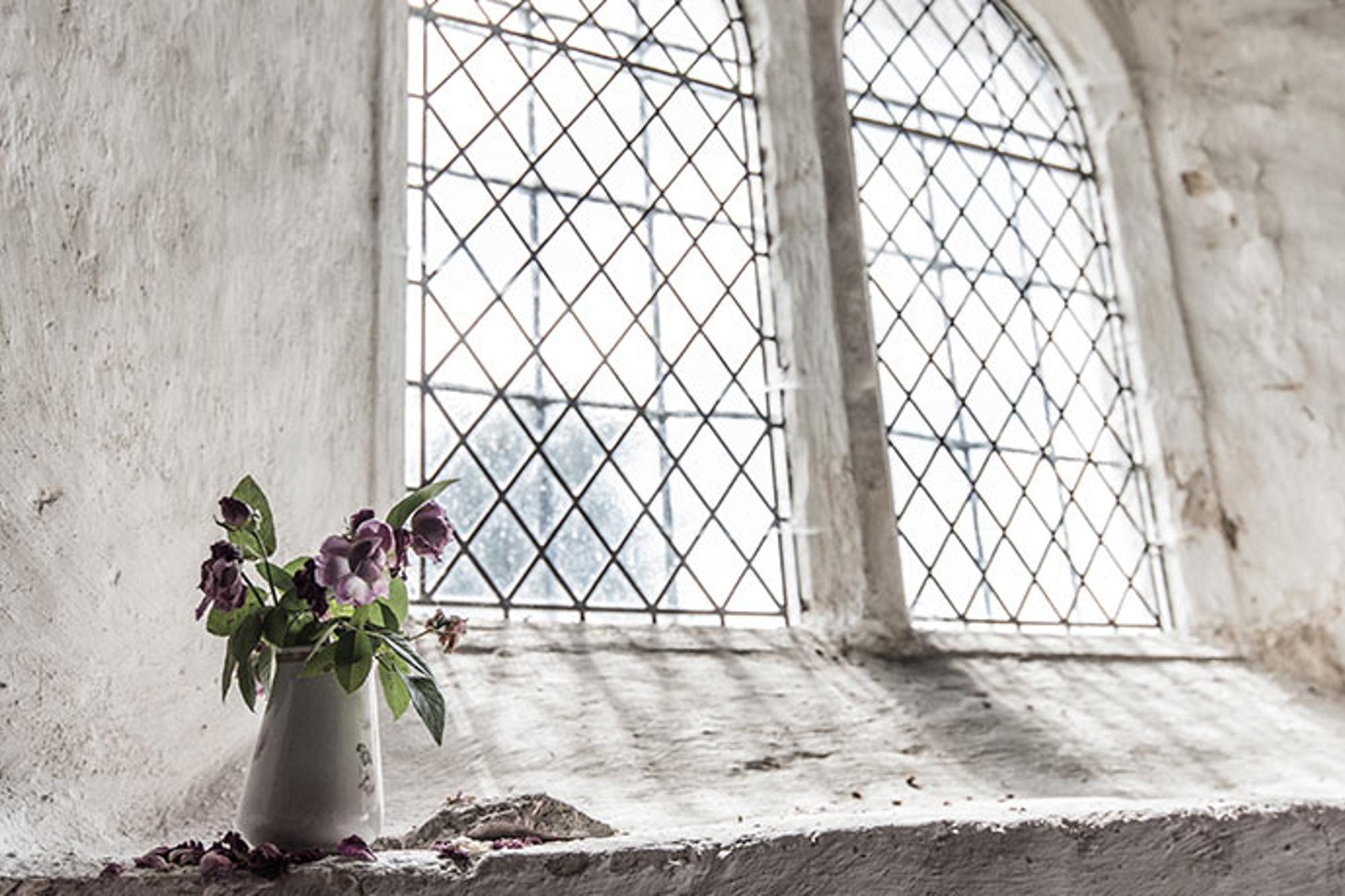 En vas med lila blommor. Vasen står vid en vägg av sten med fönster som släpper in ljuset.