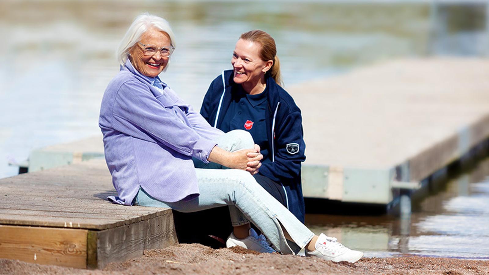 En kvinna med Frälsningsarméns sköld på sin tröja sitter på en brygga och samtalar med en äldre kvinna som tittar in i kameran och ler.