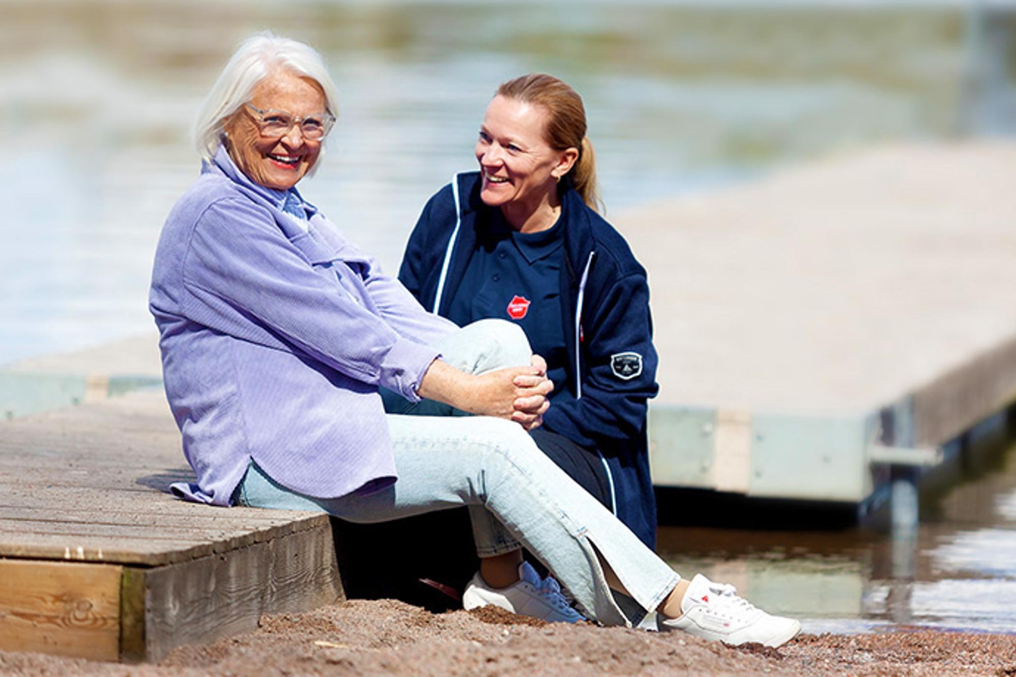 En kvinna med Frälsningsarméns sköld på sin tröja sitter på en brygga och samtalar med en äldre kvinna som tittar in i kameran och ler.
