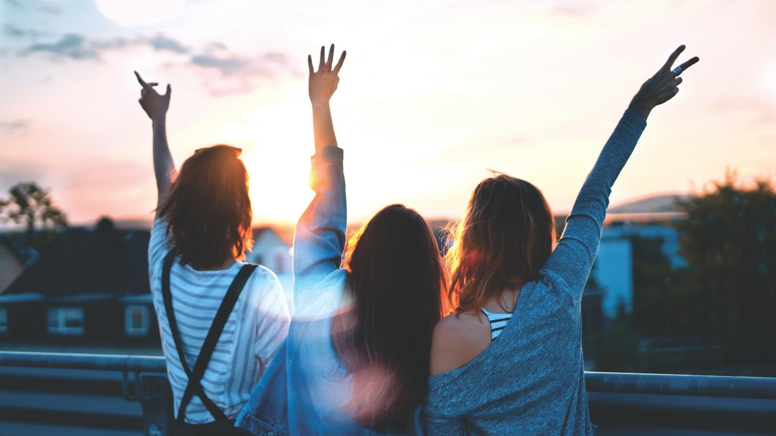 Tre unga tjejer står med ryggarna mot kameran  med händerna upp i luften mot en solnedgång