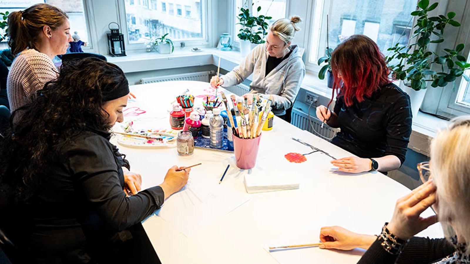 Kvinnor från en samtalsgrupp på Vårsol som sitter och målar. I mitten på bordet ligger det penslar, målarfärg och papper.