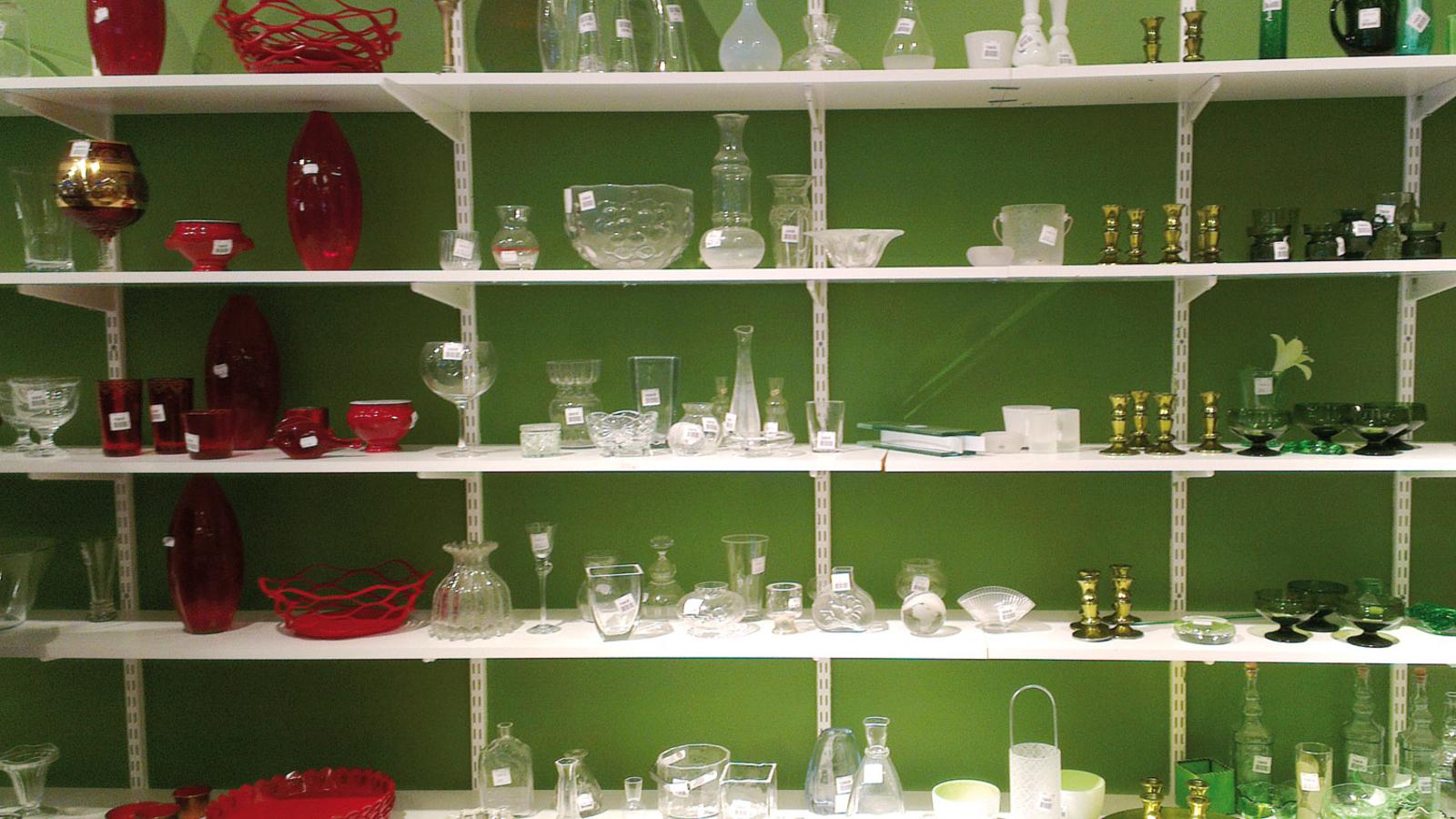 Hyllor mot en grön vägg. På hyllorna står det brickor, vaser, skålar och dekorationer i glas.