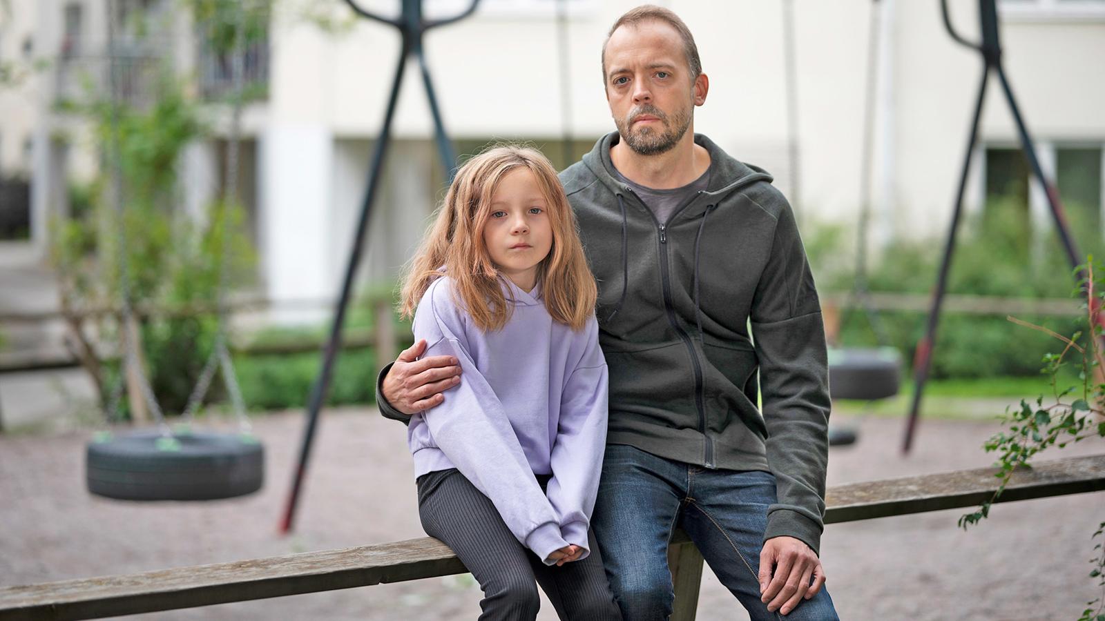 En pappa med dotter i 10-årsåldern sitter på en bank med en lekplats i bakgrunden - de ser bägge allvarliga ut.