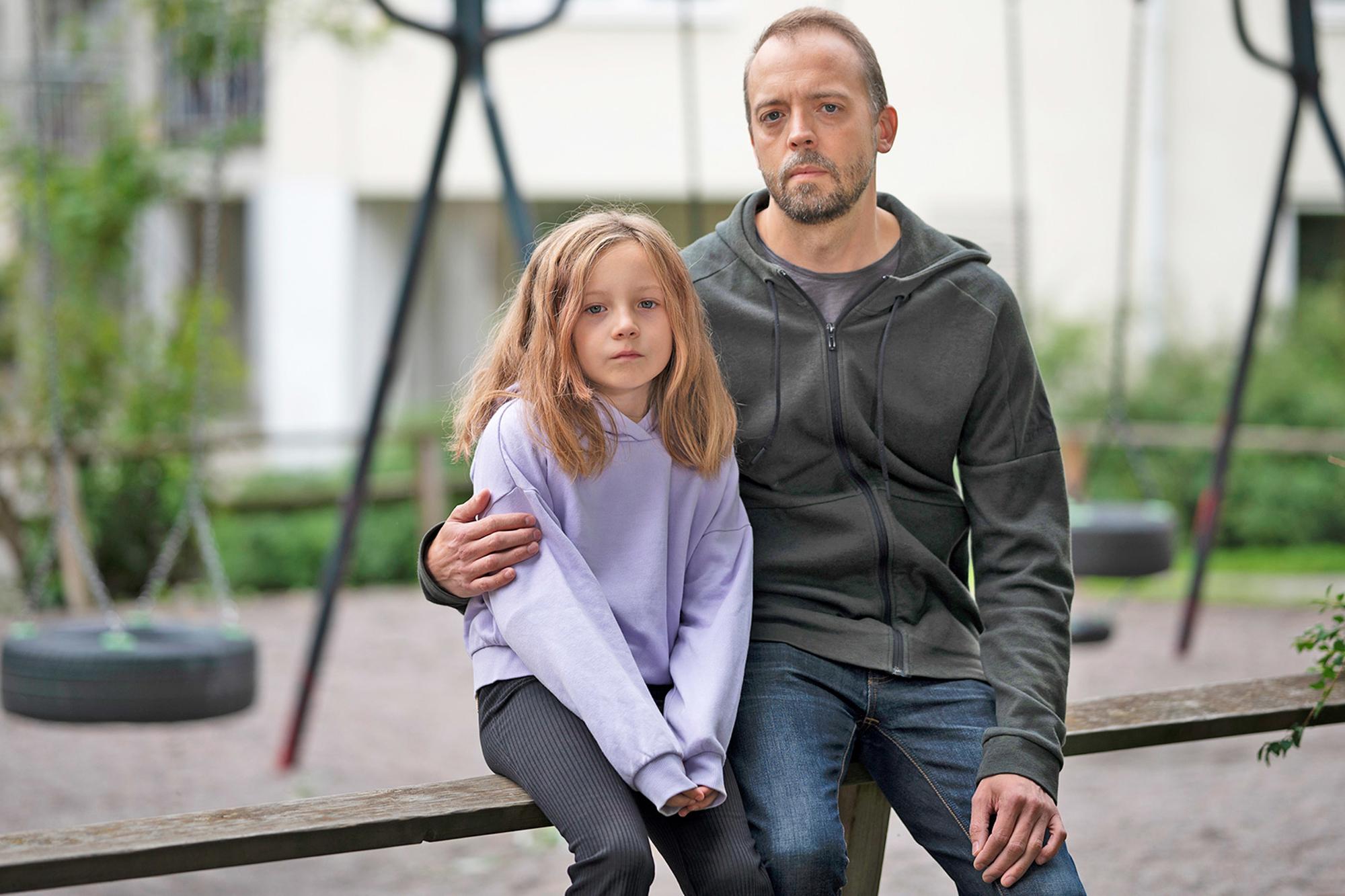 En pappa med dotter i 10-årsåldern sitter på en bank med en lekplats i bakgrunden - de ser bägge allvarliga ut.
