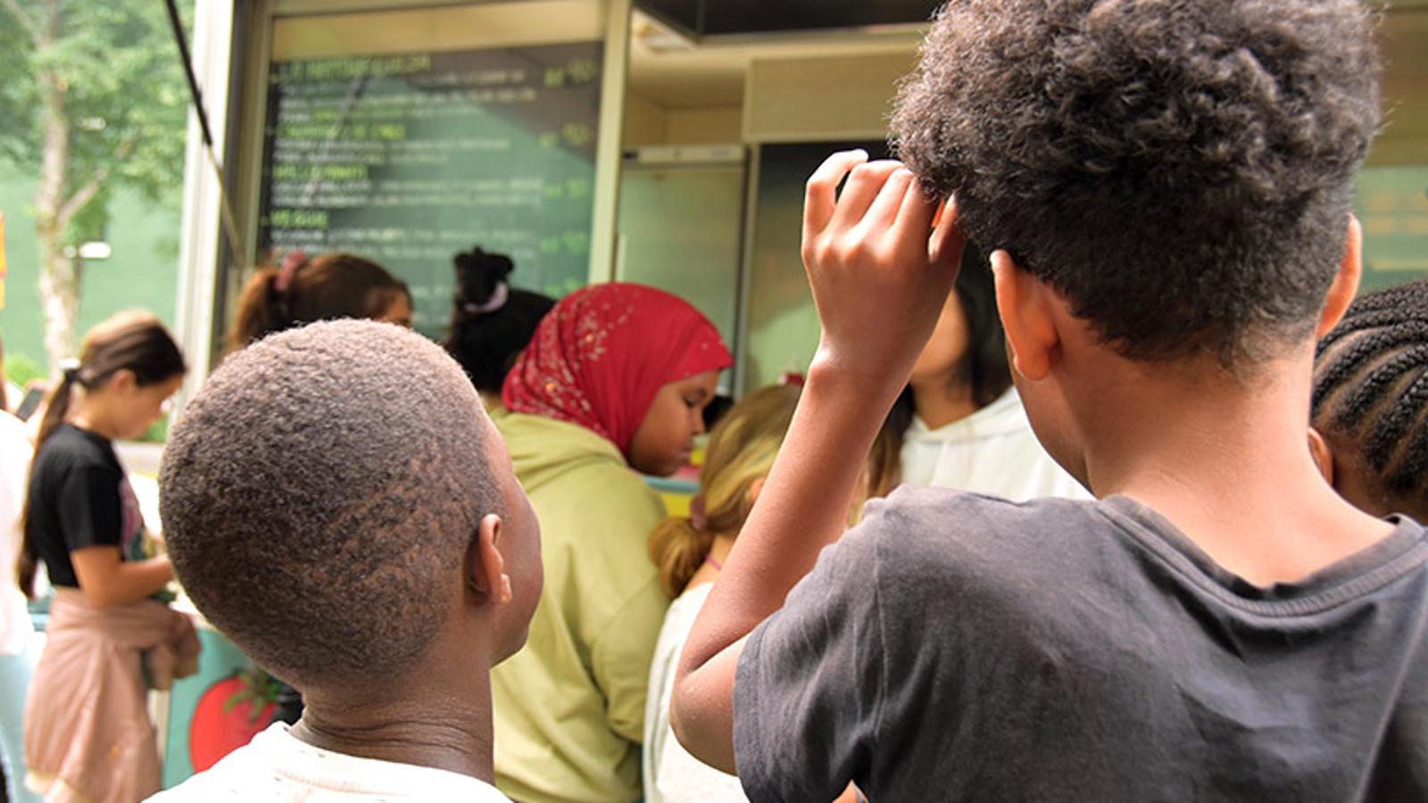 Barn med ryggtavlan mot kameran. De står i kö till en foodtruck som serverar lunch till barnen på kollot.