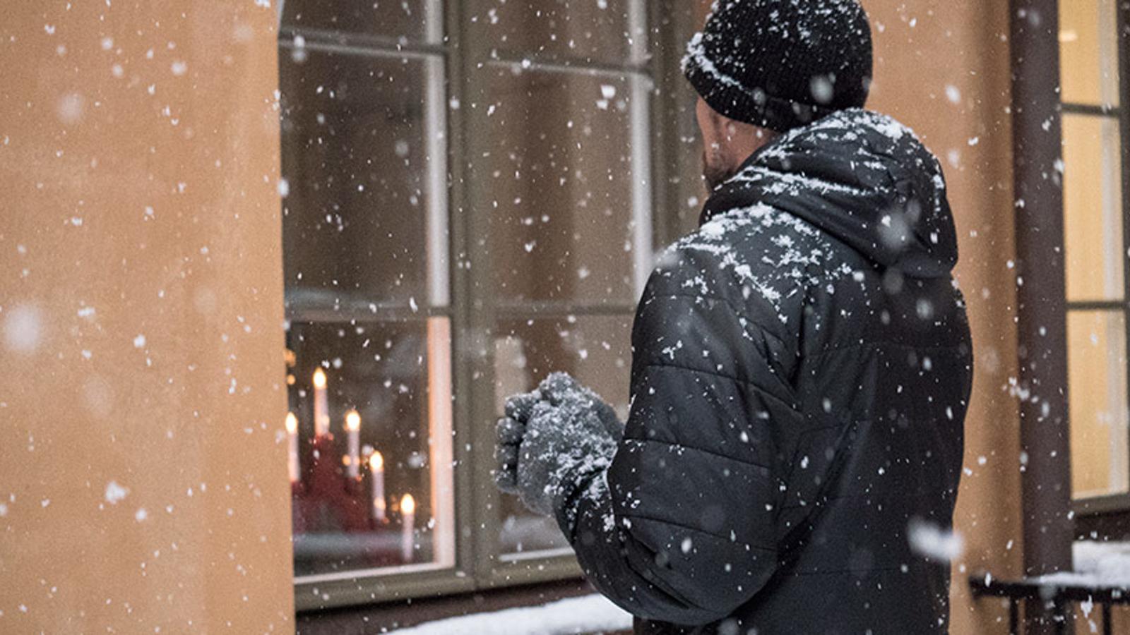 En man som står utomhus iklädd en svart jacka, gråa vantar och en svart mössa. Det snöar. Mannen står med ryggtavlan mot kameran och tittar på ett fönster med en röd adventsljusstake som lyser.
