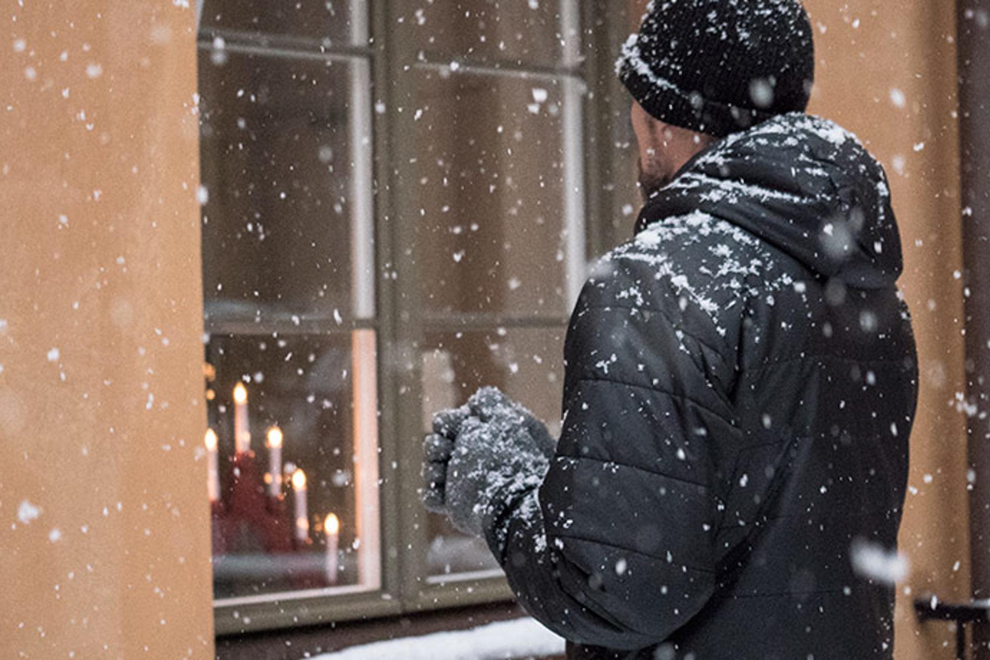 En man som står utomhus iklädd en svart jacka, gråa vantar och en svart mössa. Det snöar. Mannen står med ryggtavlan mot kameran och tittar på ett fönster med en röd adventsljusstake som lyser.