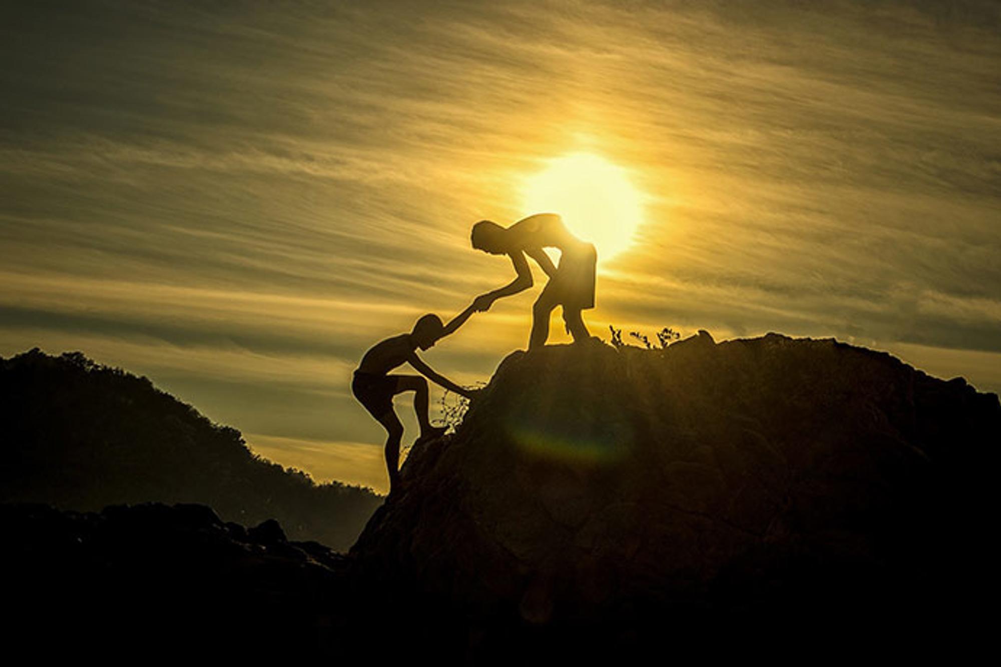 En person som får hjälp upp för ett berg av en annan person, och med en solnedgång i bakgrunden.