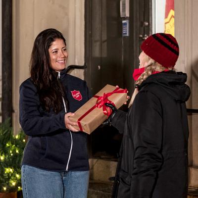 En ung kvinna med Frälsningsarméns sköld på jackan står utanför en port och räcker fram en julklapp till en kvinna.