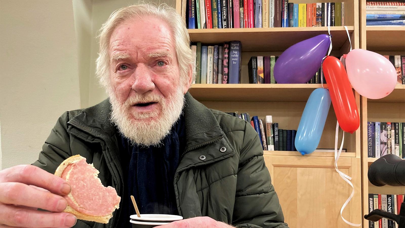 En äldre man med skägg sitter med en smörgås och en mugg i händerna - i bakgrunden syns en bokhylla och en knippe uppblåsta ballonger.