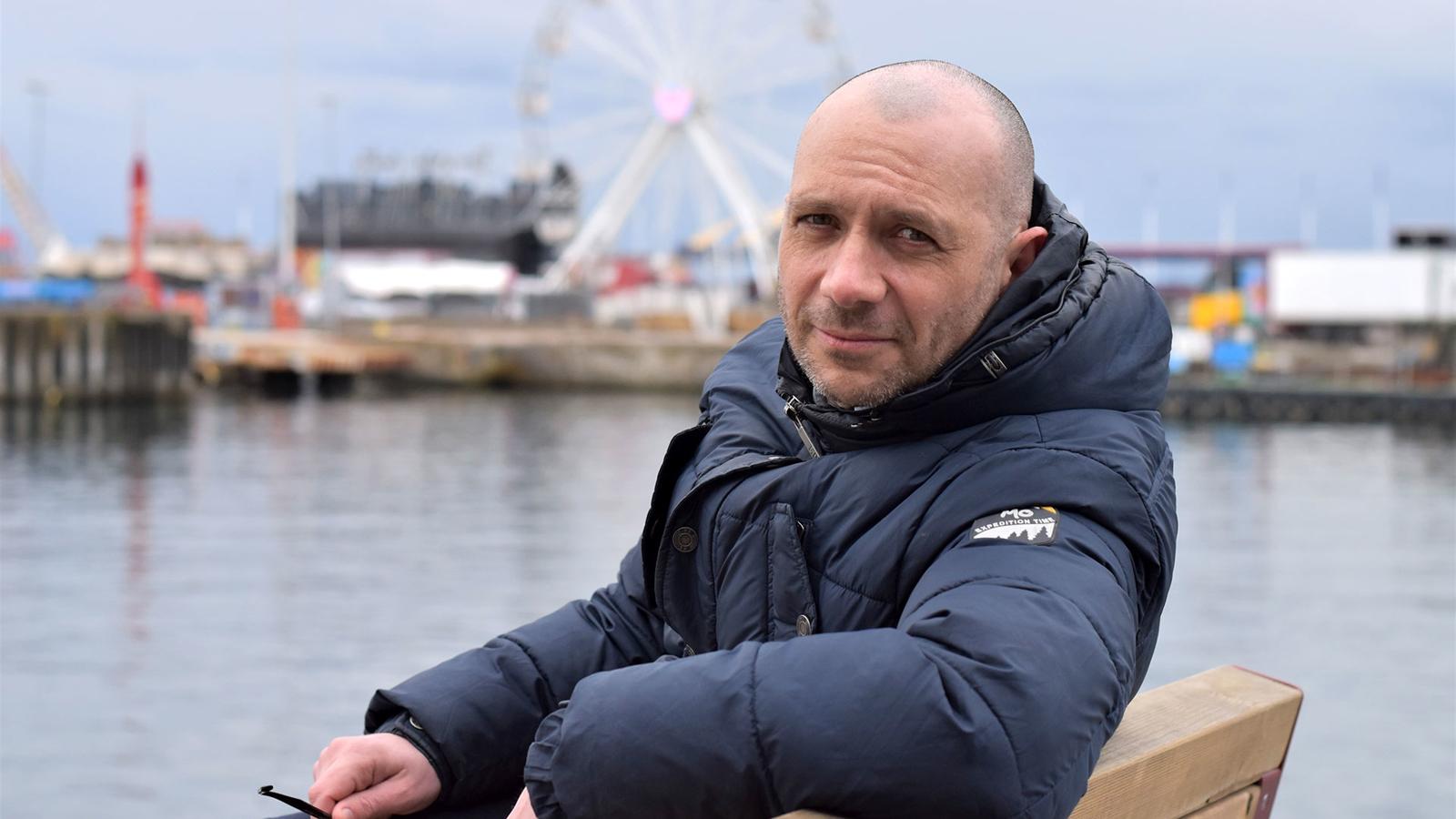 Porträttbild av Mark Cotterill. Han sitter på en bänk med blicken i kameran, och bakom honom syns en hamn.