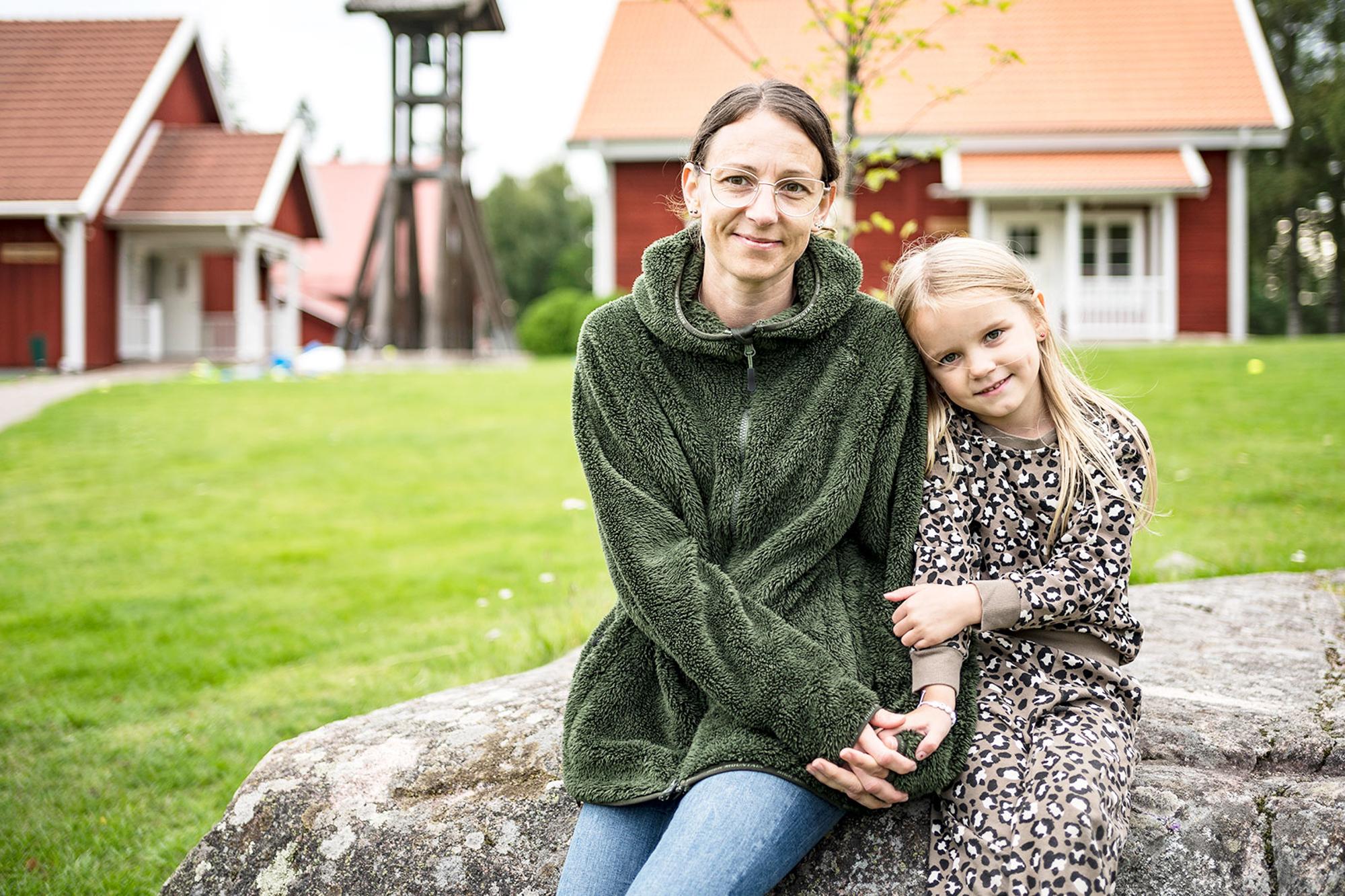 Micéla sitter med dottern Celine på en sten framför en gräsmatta och några rödmålade hus.