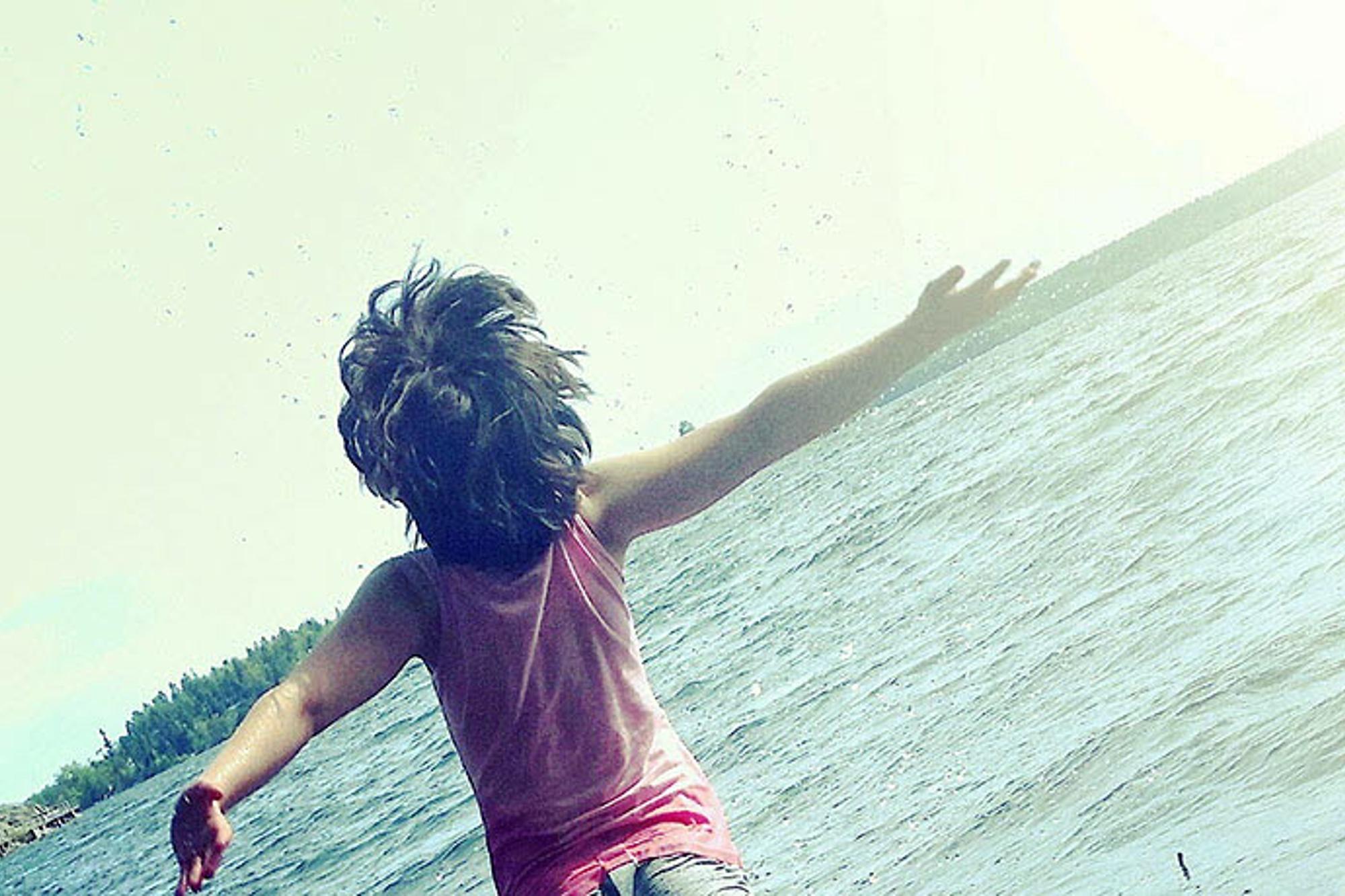 Ett barn med ryggtavlan mot kameran. Barnet står med armarna upp i luften vänd mot en sjö.