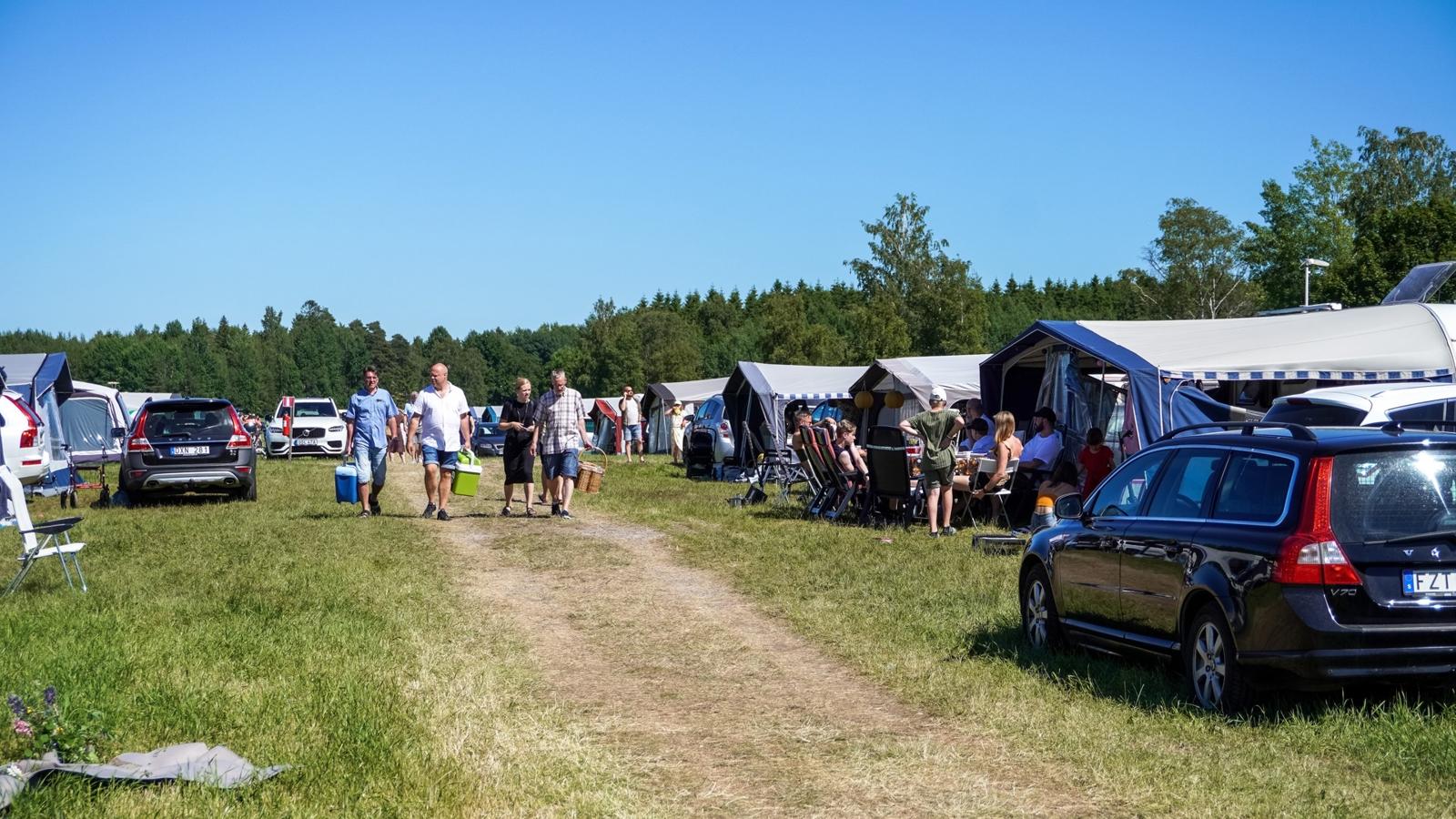 Människor går genom en campingplats med bilar och husvagnar med kylväskor i händerna.