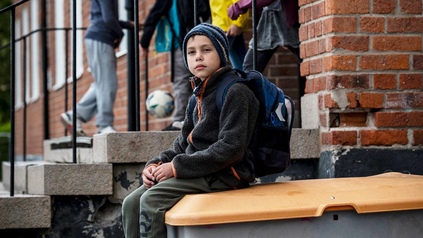 En pojke med ryggsäck sitter på en låda på en skolgård och ser uppgiven ut. Man ser andra barn i bakgrunden.