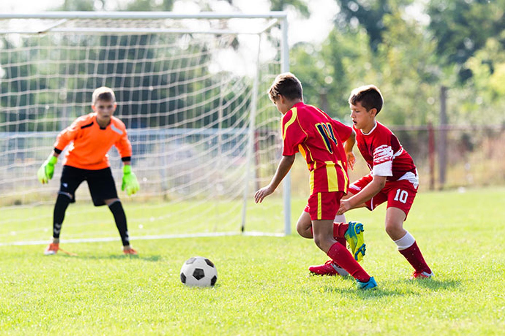 Tre pojkar som spelar fotboll på en fotbolls plan. Två av pojkarna jagar bollen och den tredje står i mål.