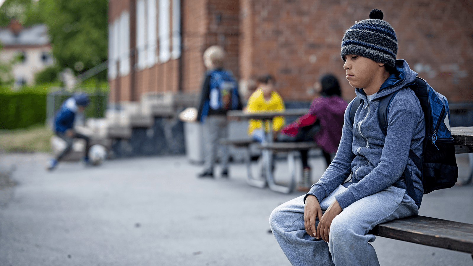En pojke med ryggsäck sitter på en bänk på en skolgård och ser uppgiven ut. Man ser andra barn i bakgrunden.