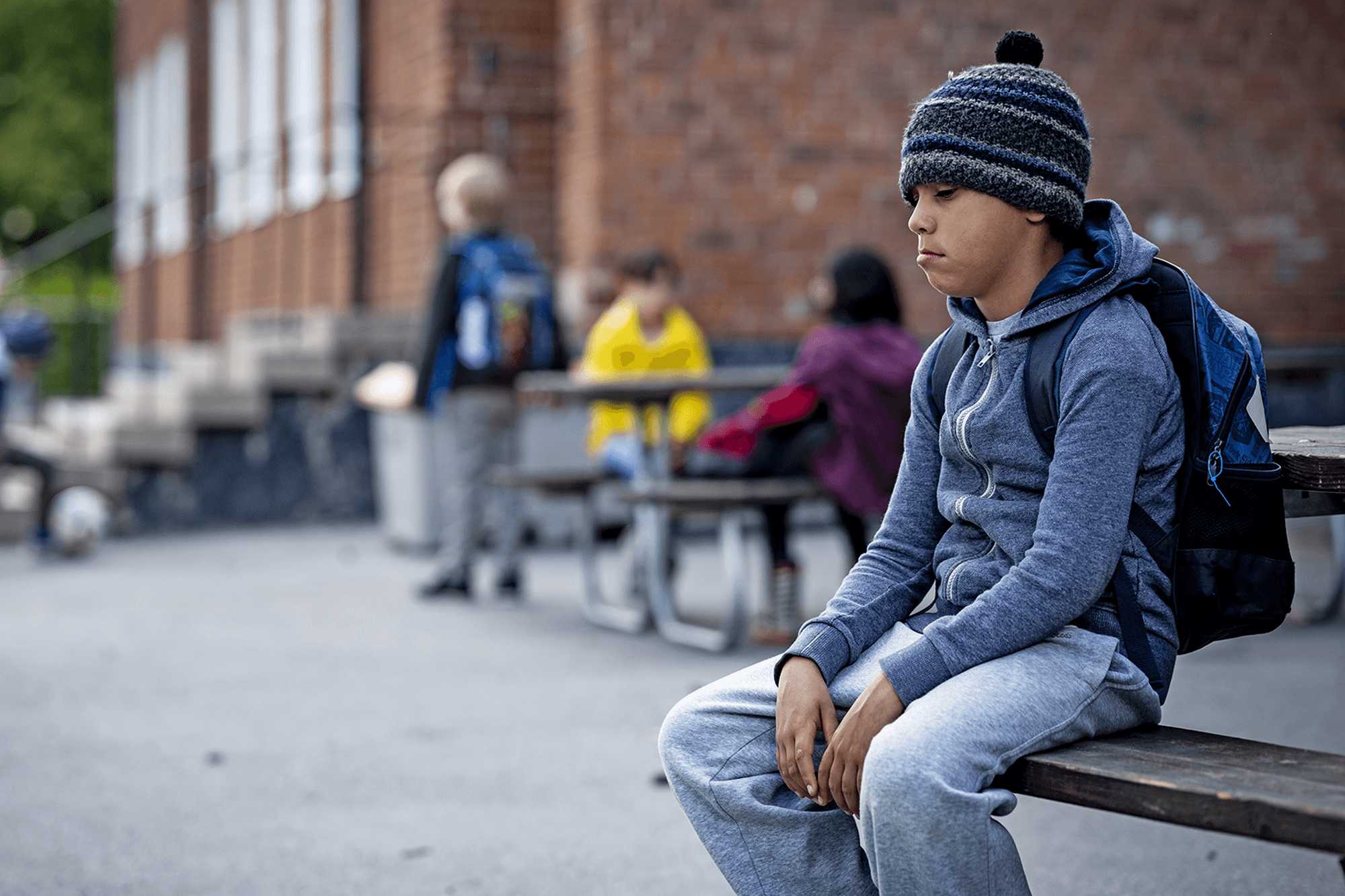 En pojke med ryggsäck sitter på en bänk på en skolgård och ser uppgiven ut. Man ser andra barn i bakgrunden.