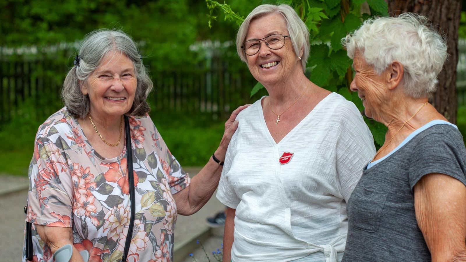 Två leende äldre kvinnor står utomhus tillsammans med en kvinna som har Frälsningsarméns sköld på tröjan.