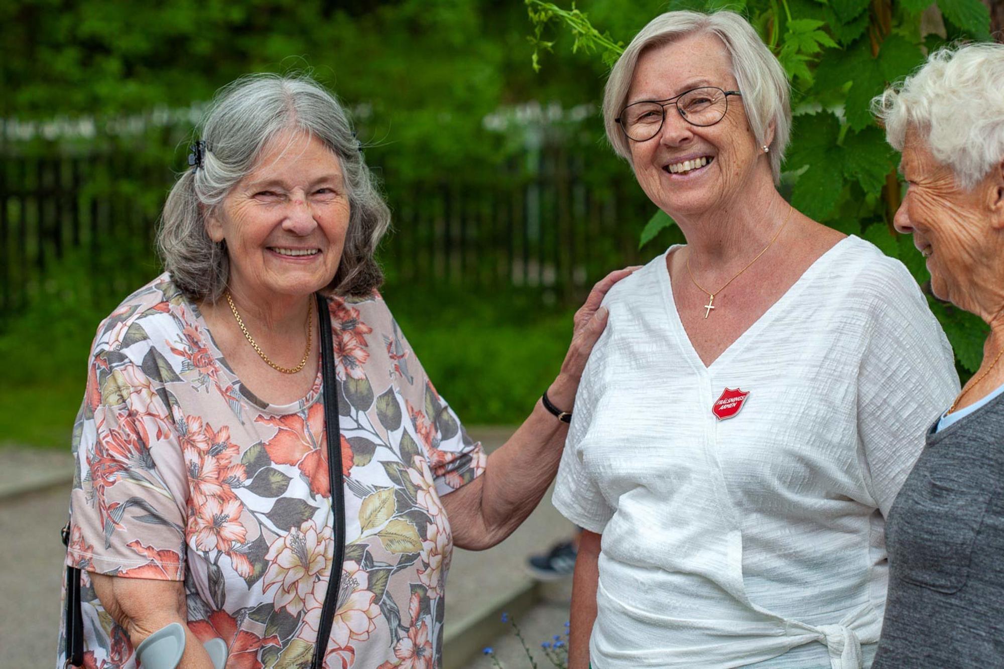 Två leende äldre kvinnor står utomhus tillsammans med en kvinna som har Frälsningsarméns sköld på tröjan.