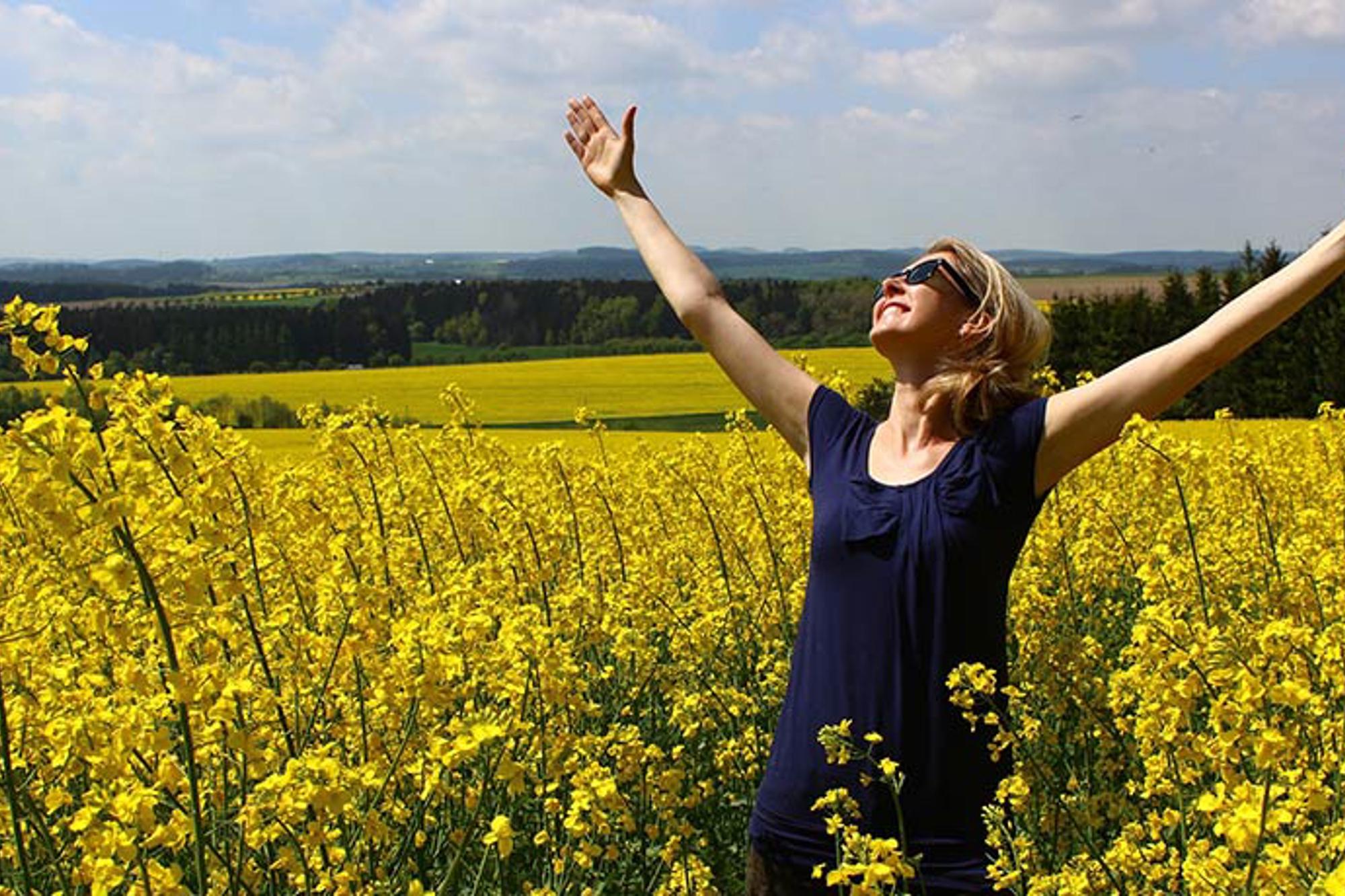En glad kvinna på ett fält av gula blommor. Hon sträcker båda armarna upp i luften och tittar upp mot himlen.