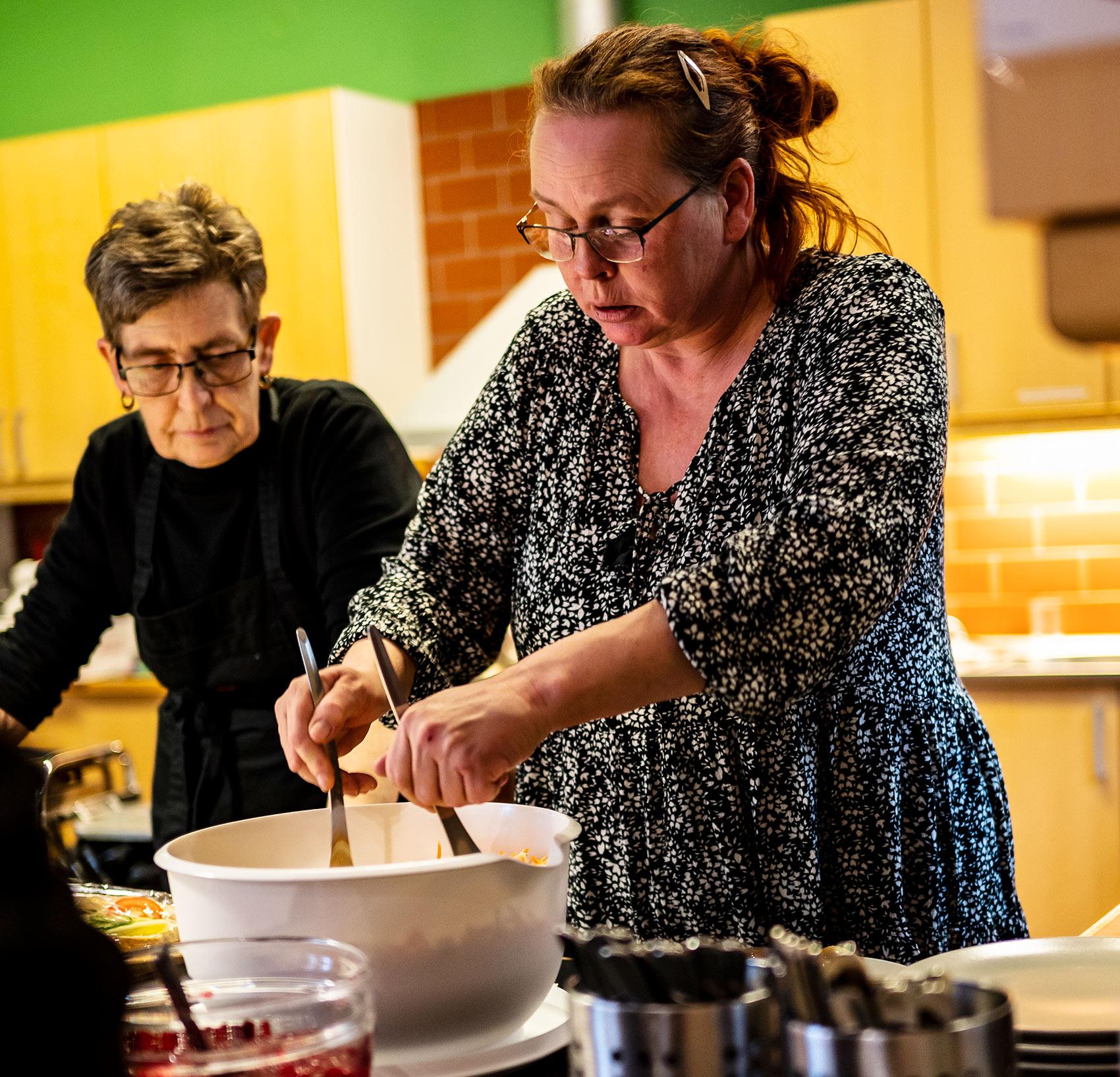 Eva Andersson som står i köket på kåren i Ystad. Hon gör iordning maträtter som skall serveras, och till vänster om henne står en kollega.
