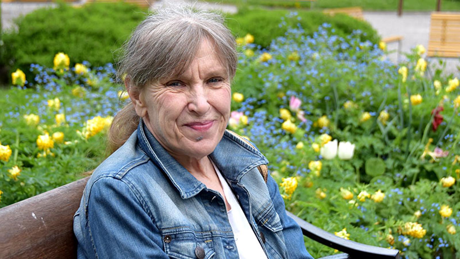 Porträttbild av Maud Bäck. Hon sitter på en bänk och ler mot kameran. I bakgrunden syns buskar och blommor.