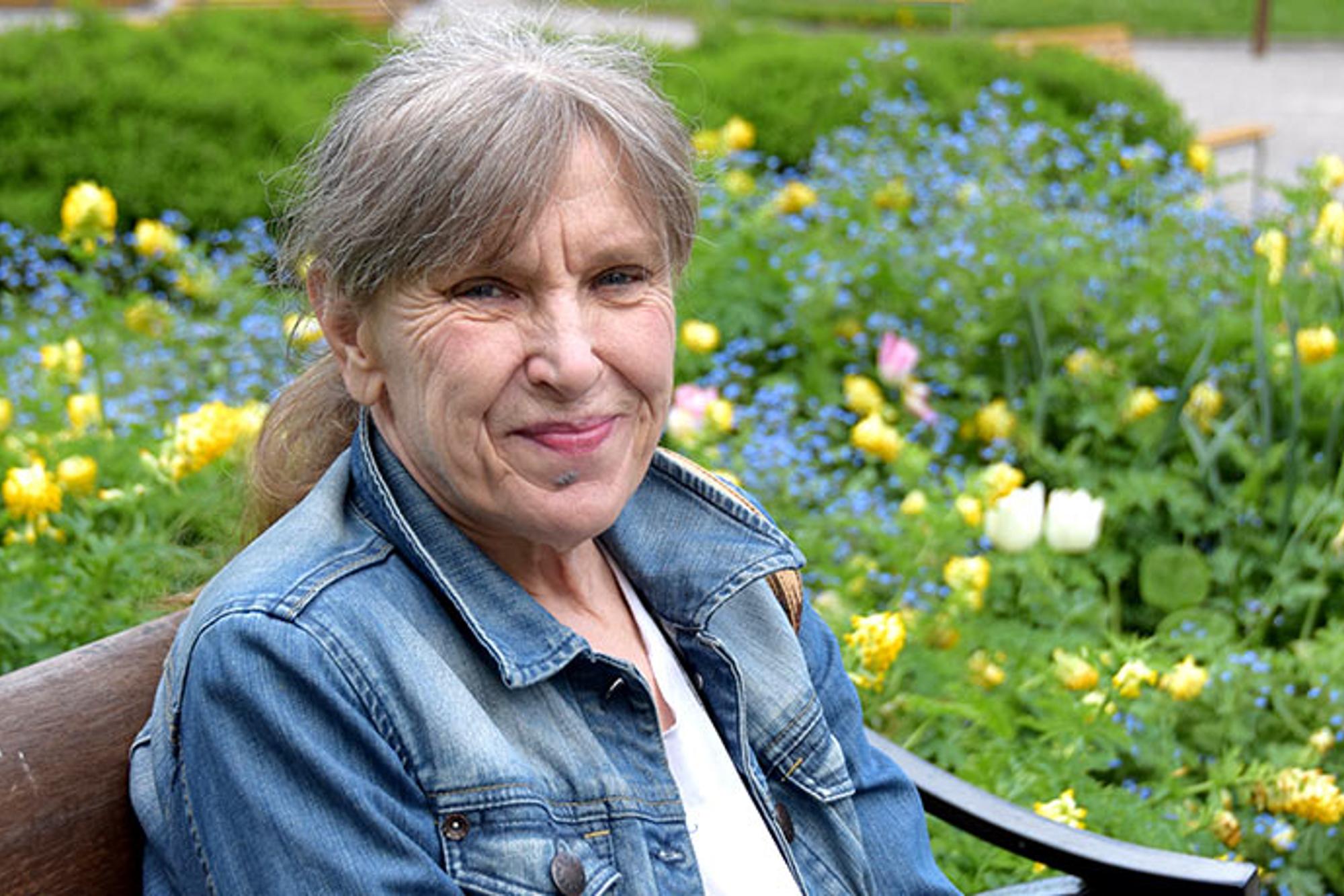Porträttbild av Maud Bäck. Hon sitter på en bänk och ler mot kameran. I bakgrunden syns buskar och blommor.