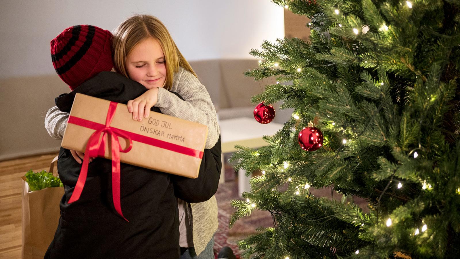 En mamma kramar om sin dotter bredvid en julgran, flickan håller i ett paket och en kasse mat står vid deras sida. 