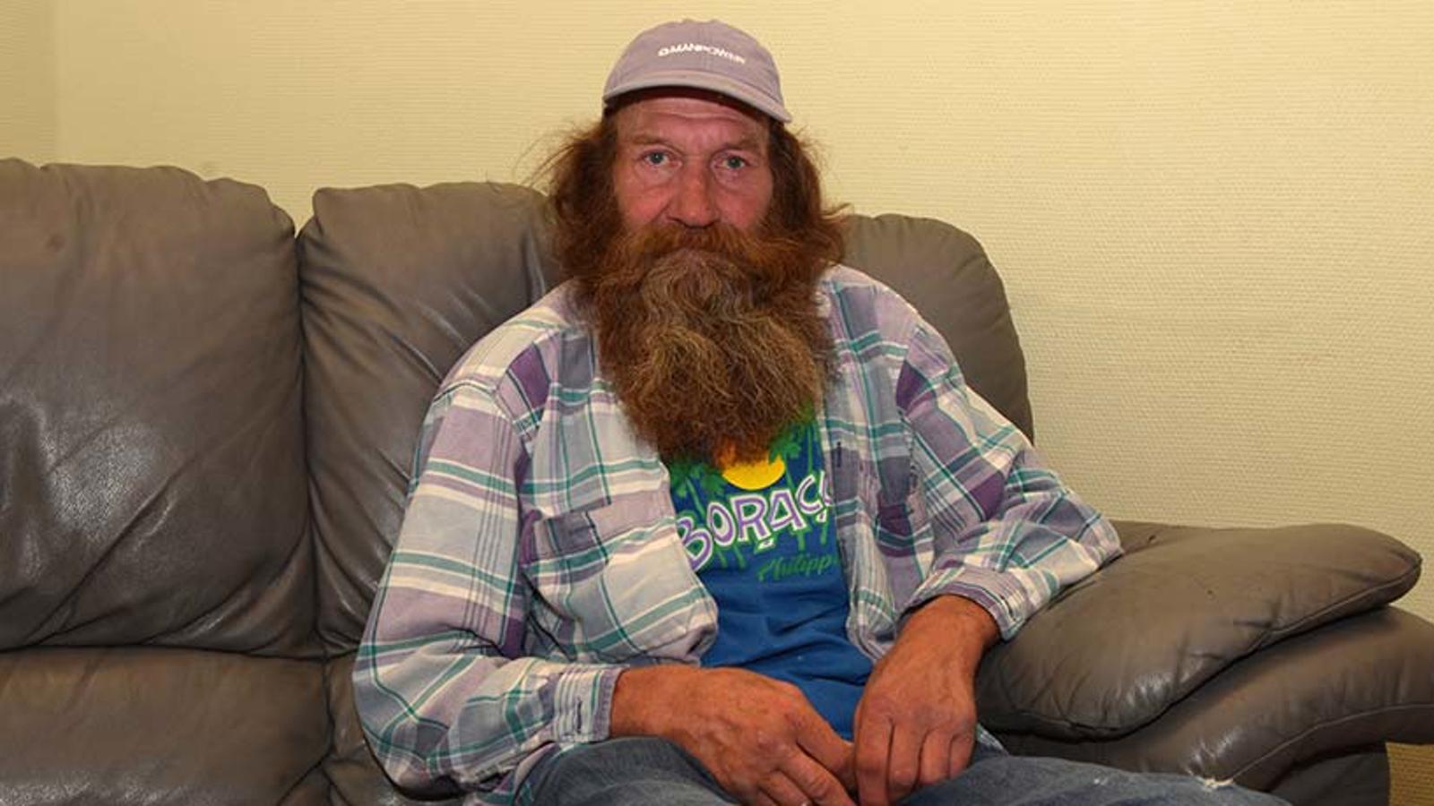 En äldre man med skägg, Kenneth, som sitter i en soffa och tittar in i kameran. Han är iklädd en blåa tröja med tryck och över tröjan har han en rutig skjorta.